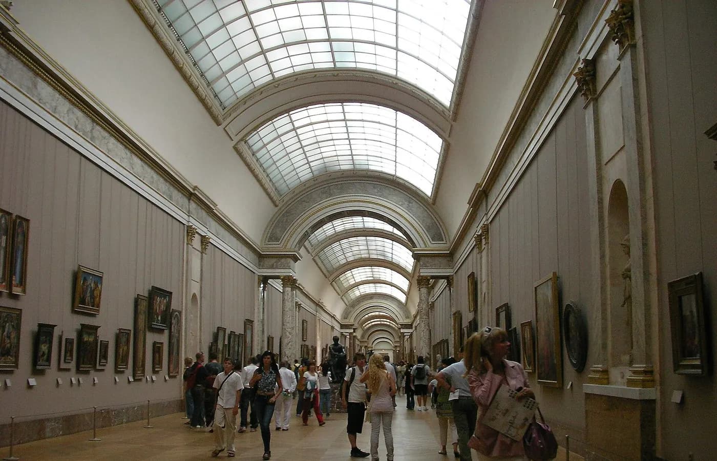 Grande galerie d’un musée, bordée de tableaux, parcourue par des visiteurs.